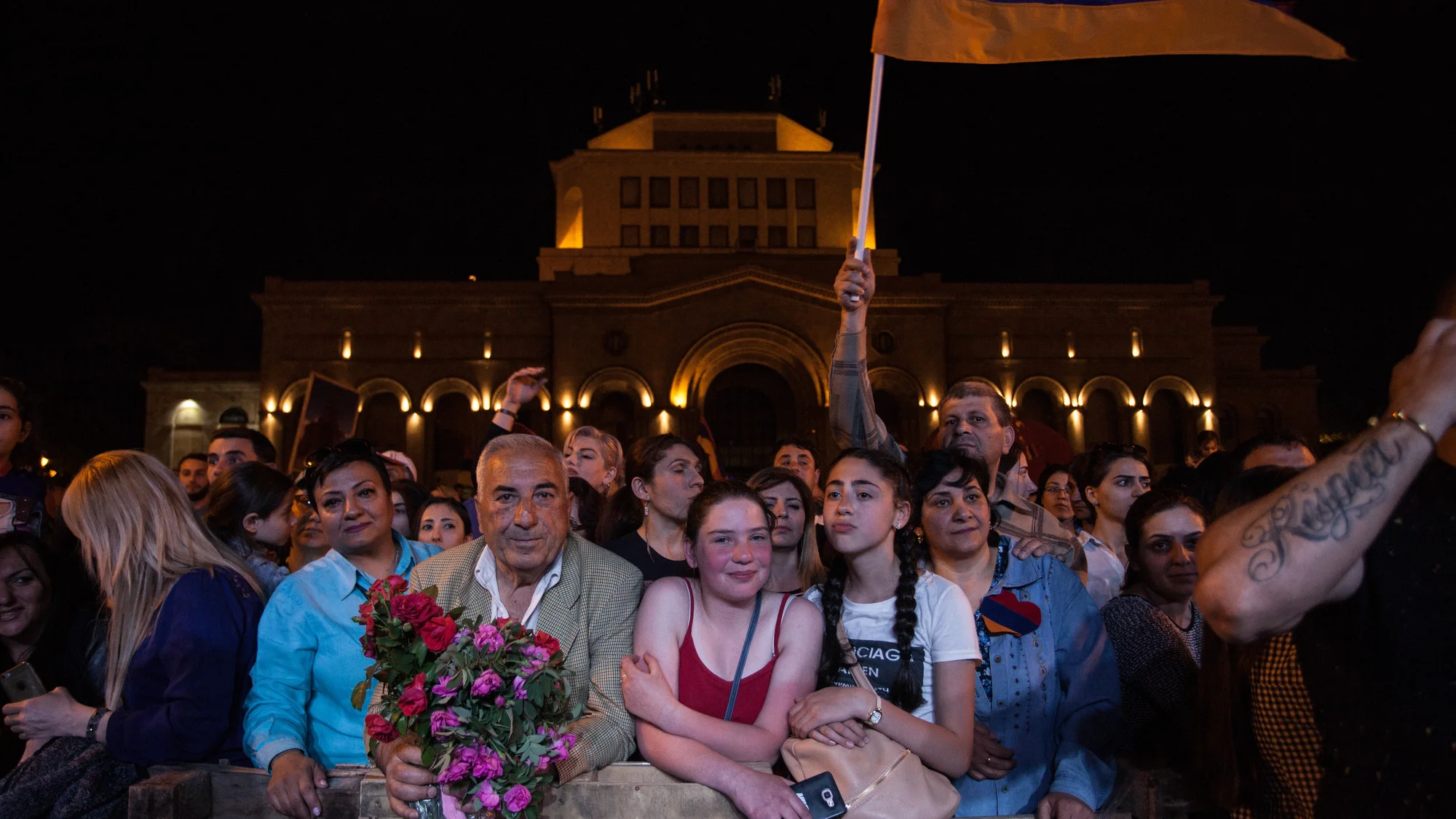 A crowd gathers at night in Yerevan during the 2018 Armenian Revolution, with flowers and a flag raised above the group.
