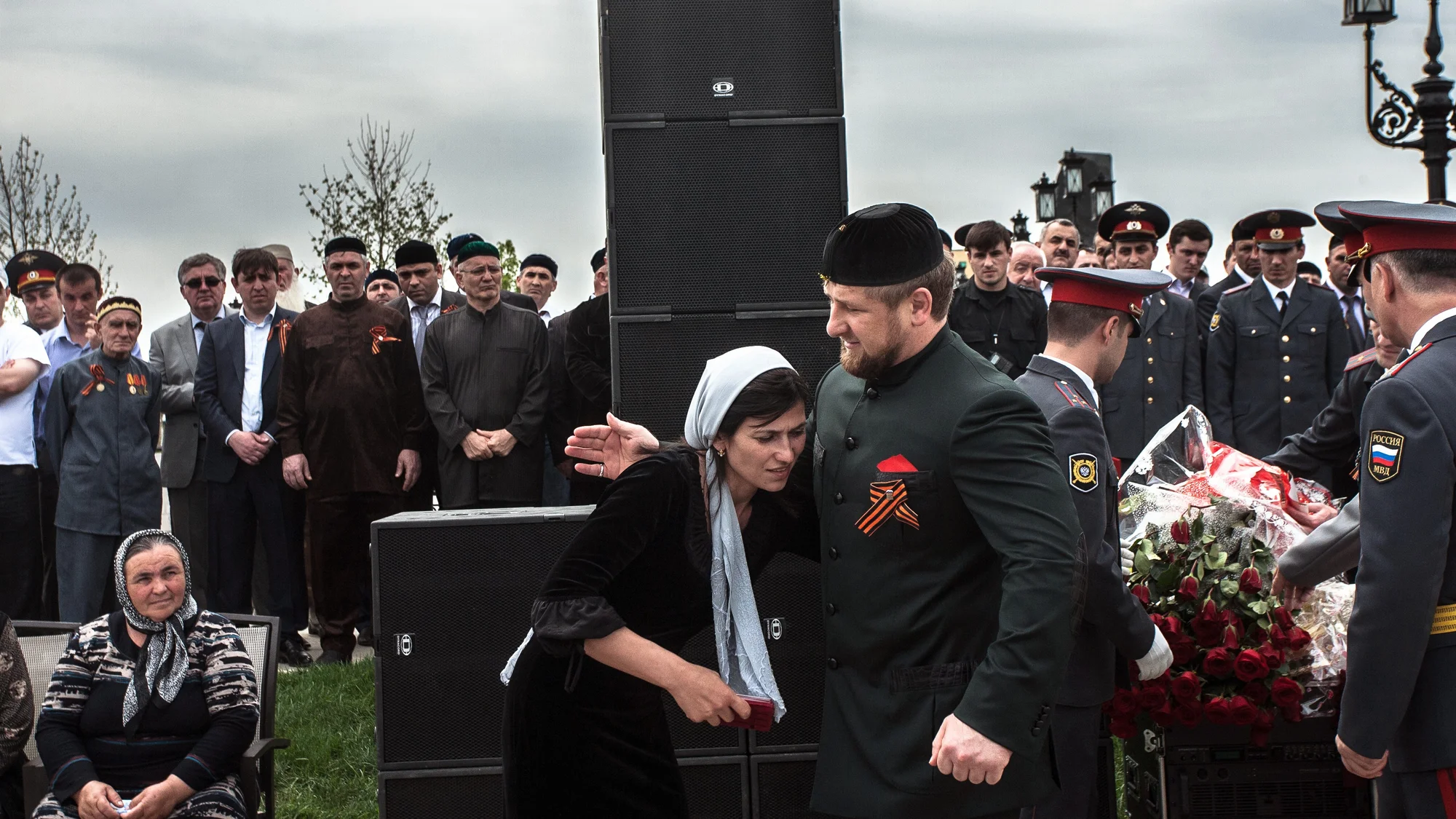 A woman in a headscarf stands beside Ramzan Kadyrov, surrounded by a crowd and uniformed officers in Chechnya.