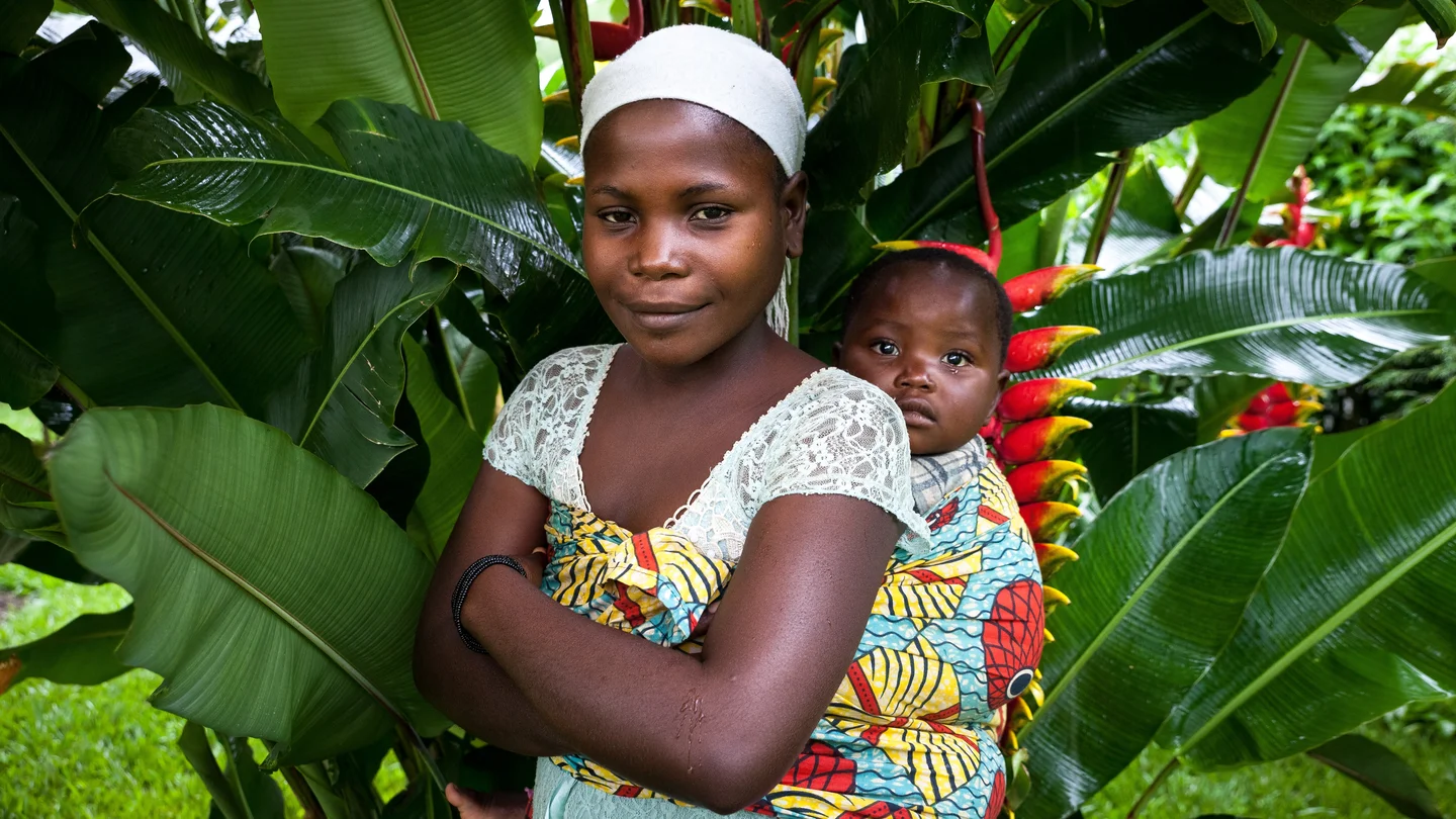 A woman in East Kivu carries a young child on her back amid lush green foliage.