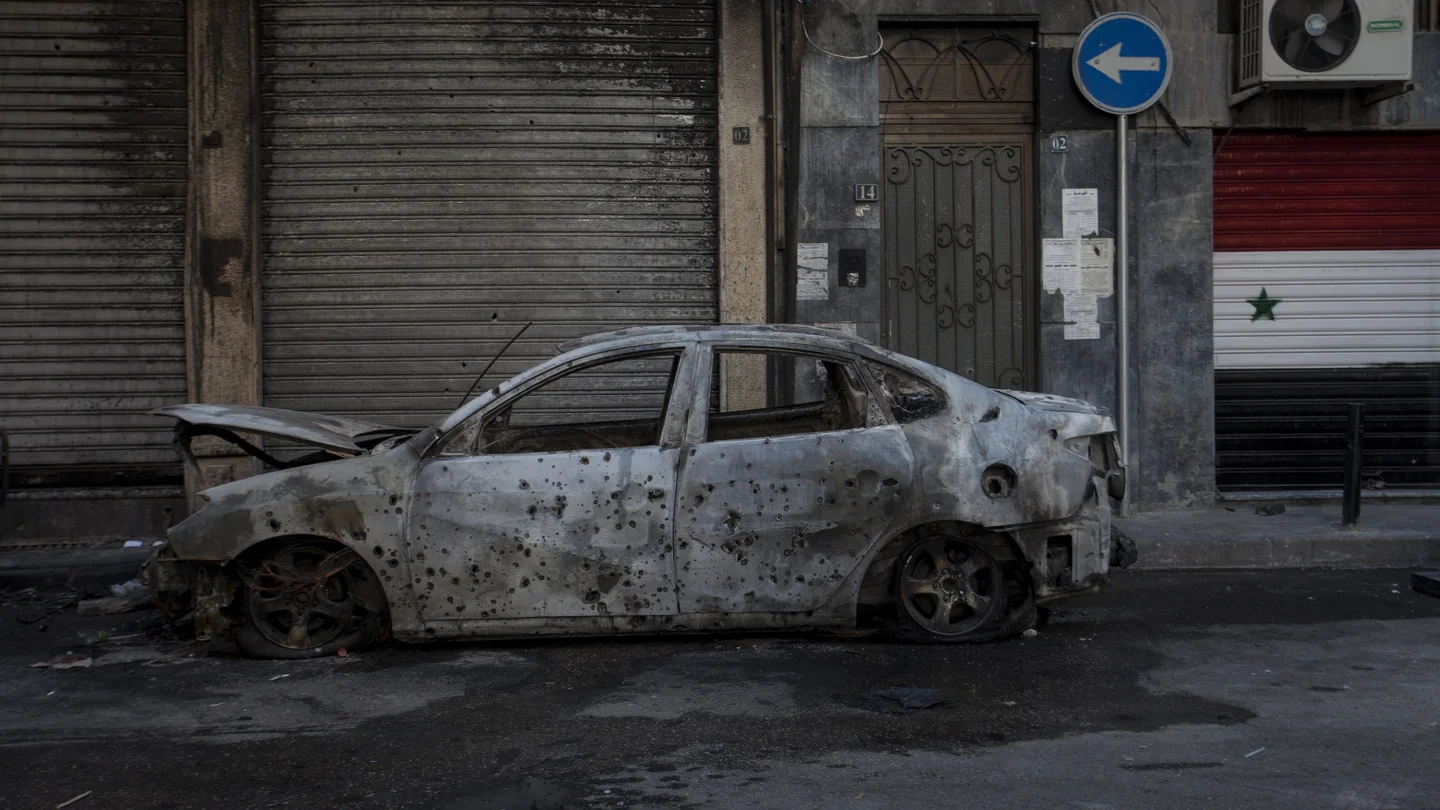 A burned-out car on a street in Damascus, beside shuttered buildings during the Syrian civil war.