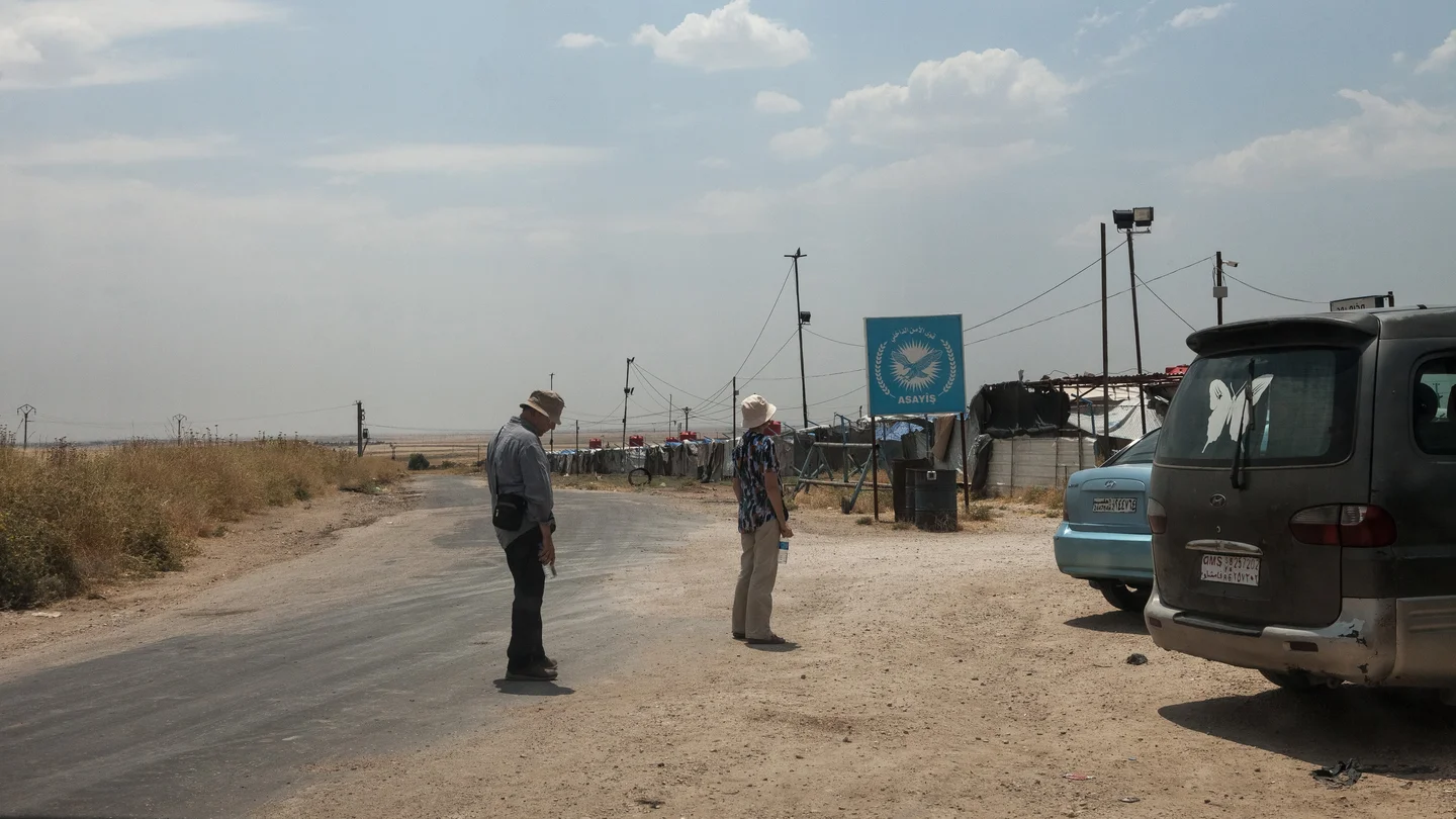 Mr. and Mrs. Lopez, a retired French couple looking for their grandchildren raised by former jihadists, stand on a dusty roadside near a checkpoint in Syrian Kurdistan.