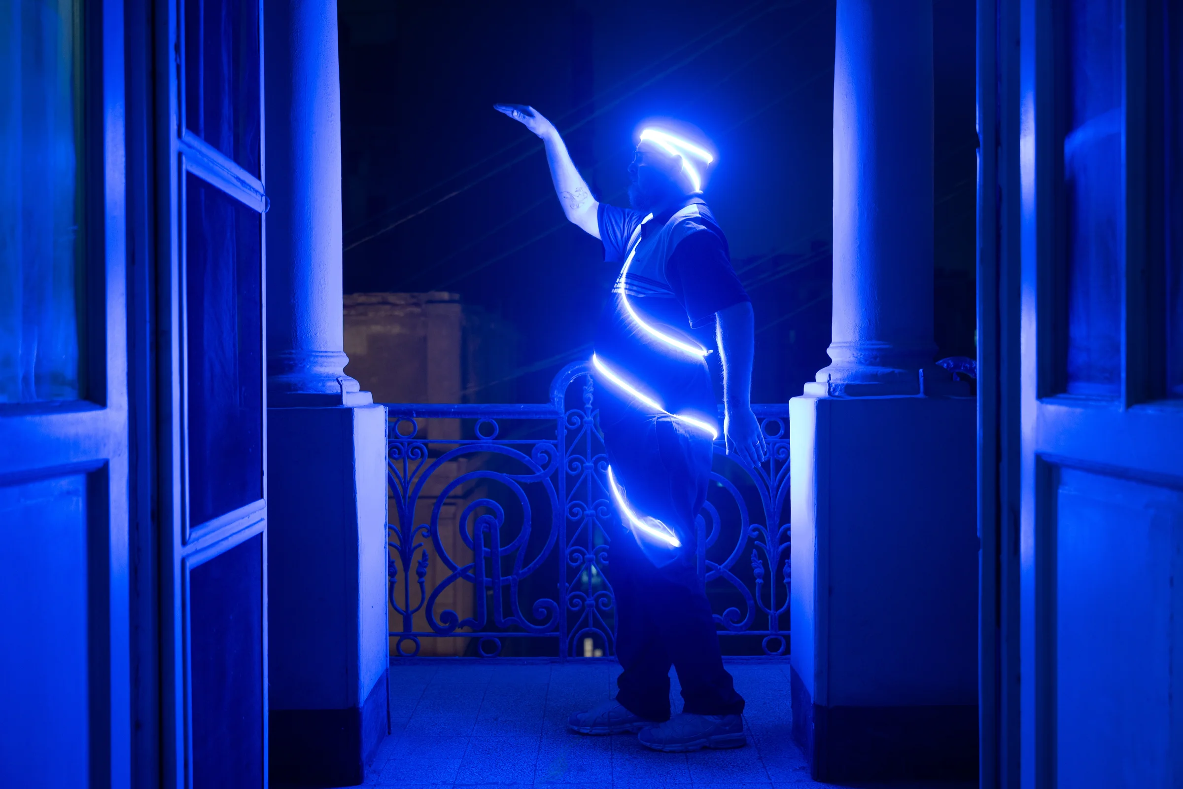 A person wrapped in blue light raises an arm on a balcony at night.