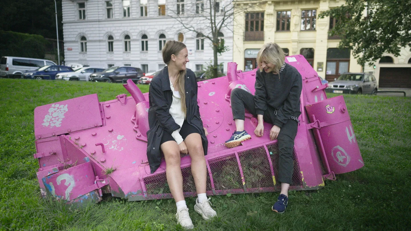 Anni and Olga smile while sitting on a pink-painted tank in a grassy square.