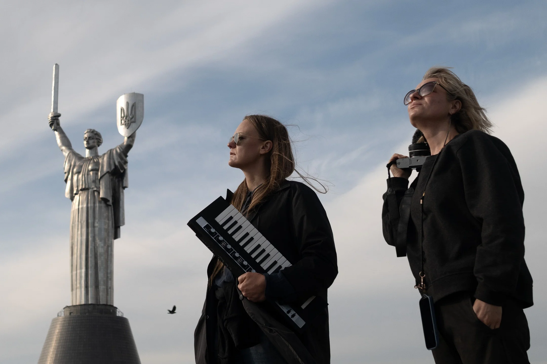 Anni Pohto and Olga Kravets, holding an electric piano and a camera, stand next to the Mother Ukraine statue in downtown Kyiv