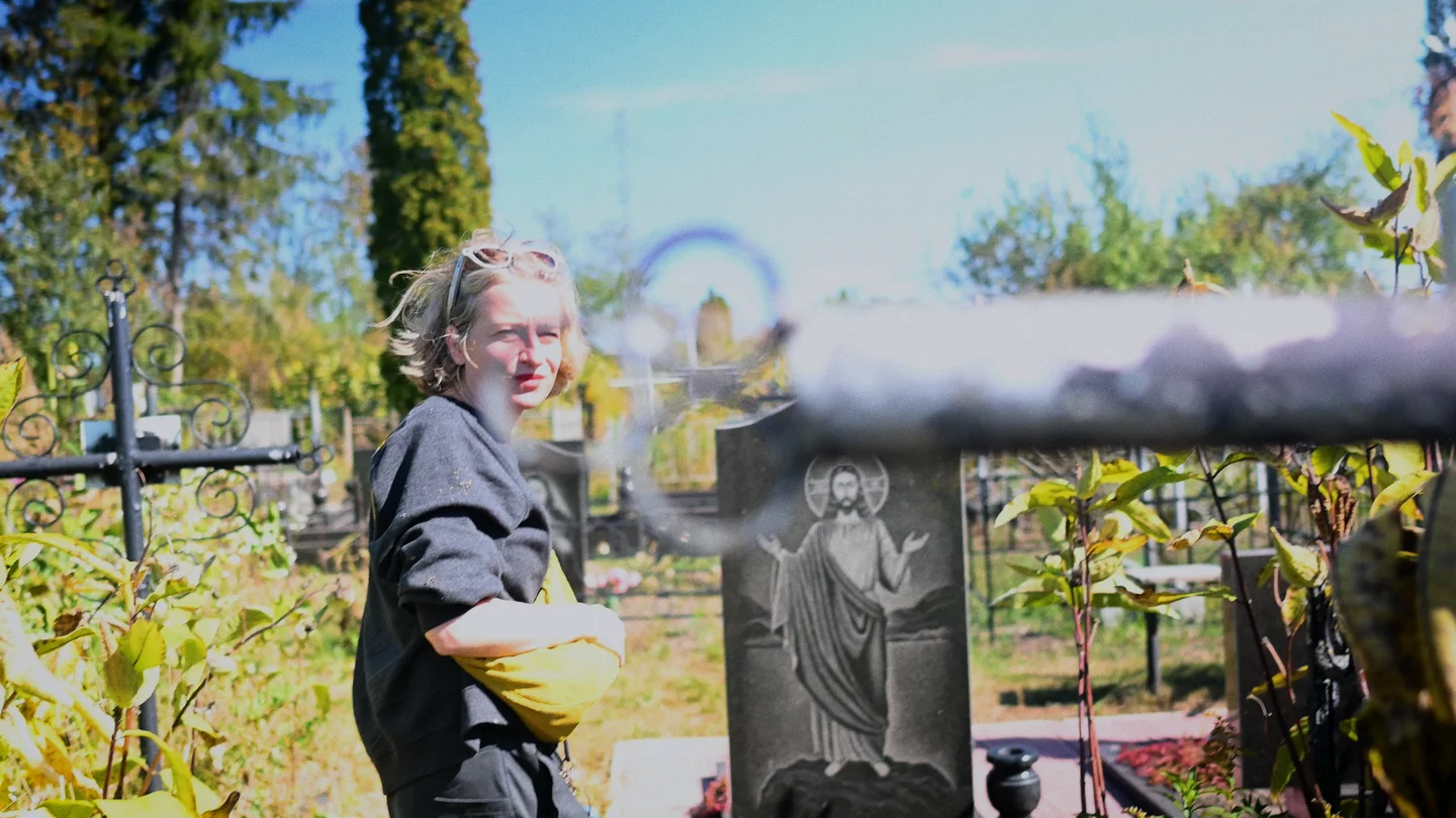 A woman with a yellow waist bag stands among gravestones in a sunlit cemetery.