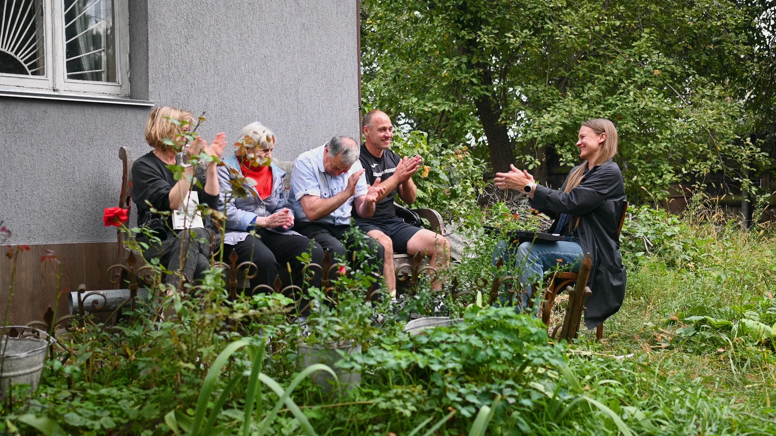 Five people sit in a garden beside a house, smiling and applauding.