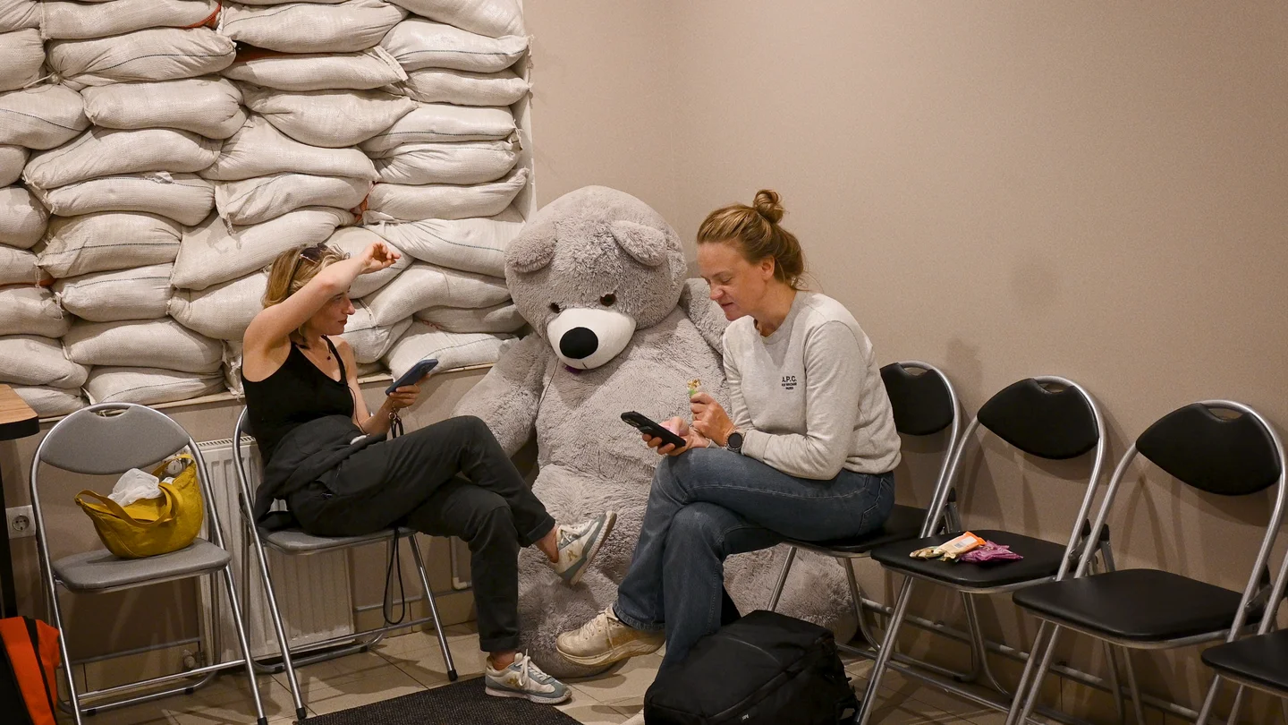 Olga Kravets and Anni Pohto wait in a bomb shelter a large teddy bear and a wall of sandbags.