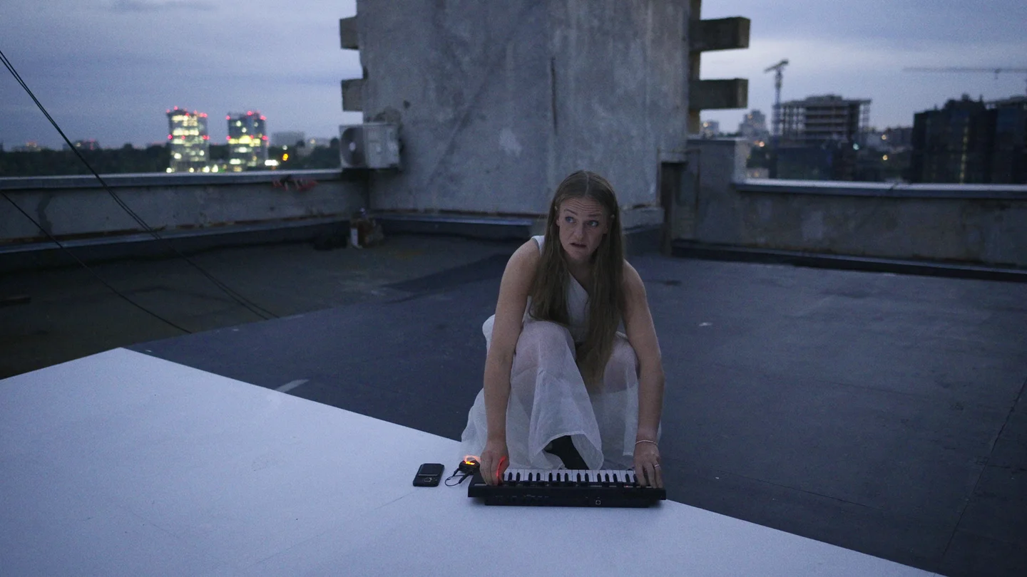 Anni Pohto in a white dress plays a small keyboard on a rooftop at dusk.